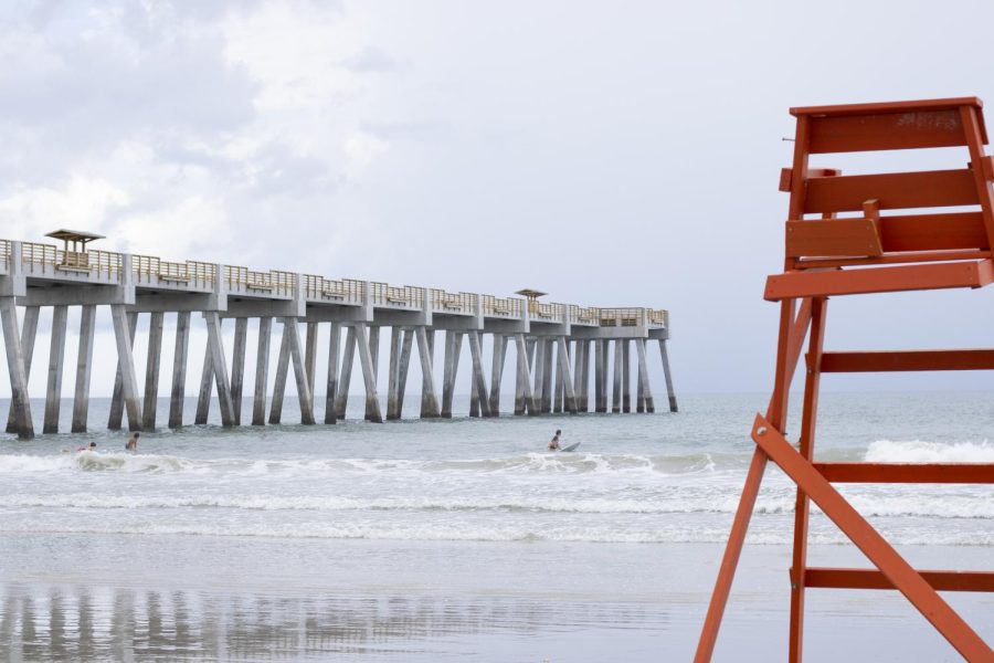 A red lifeguard chair stands on a beach as surfers and swimmers ride waves next to the newly constructed Jacksonville Beach pier