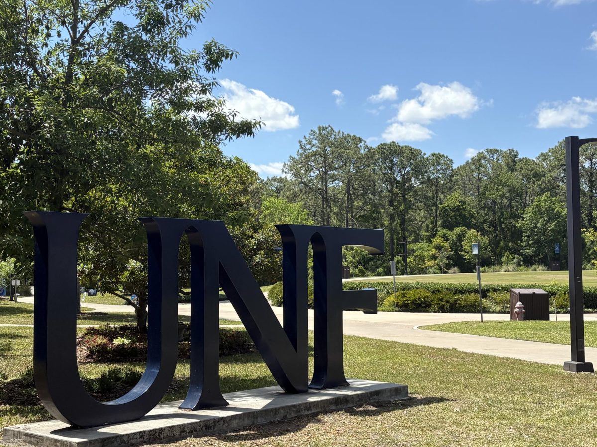A sign with large, dark blue letters spells "UNF" on a concrete stand in the grassy area next to a sidewalk. The sky is bright blue with some small white clouds and there are lots of trees in the background.
